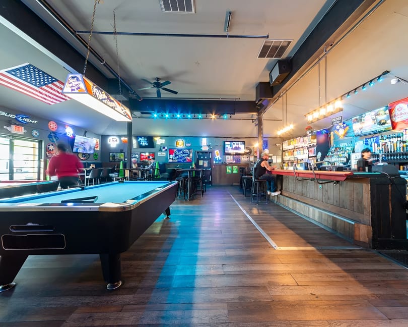 A lively bar interior with a pool table, patrons, and a well-stocked bar with neon signs.